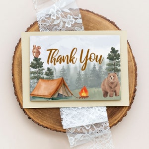 May include: A thank you card with a watercolor illustration of a forest scene with a tent, a campfire, a bear, and a squirrel. The text "Thank You" is written in brown letters.