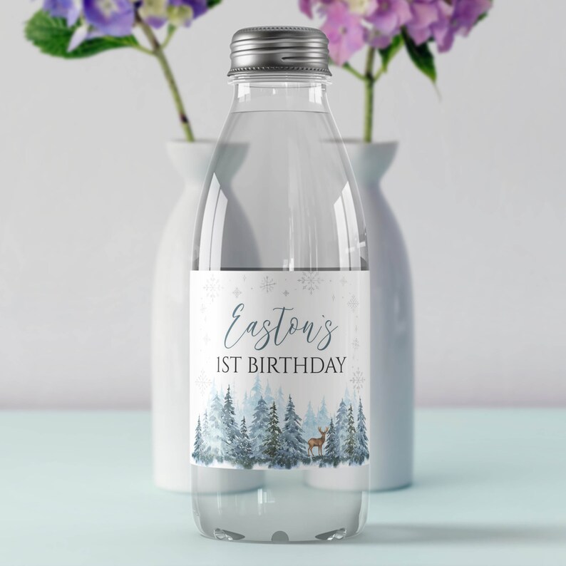 May include: A clear plastic water bottle with a silver screw cap featuring a white label. The label displays the text Easton's 1ST BIRTHDAY in blue and black font, decorated with blue pine trees, snowflakes, and a small brown deer illustration.