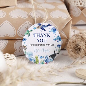 Pu&ograve; includere: Un'etichetta regalo rotonda bianca con illustrazioni ad acquerello di varie creature marine, tra cui una balena, uno squalo, un'orca, una tartaruga marina e una medusa. Il testo recita THANK YOU for celebrating with us! Love, Huson in blu e nero.