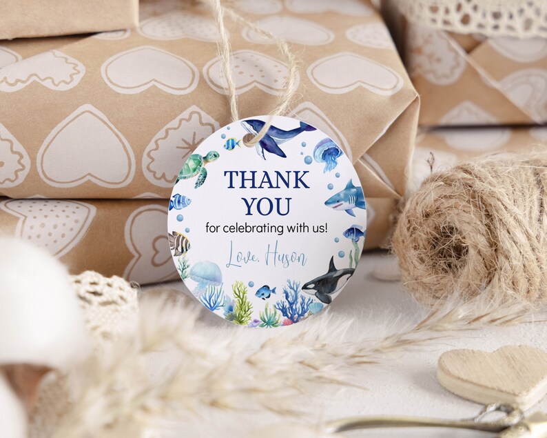 Pu&ograve; includere: Un'etichetta regalo rotonda bianca con illustrazioni ad acquerello di varie creature marine, tra cui una balena, uno squalo, un'orca, una tartaruga marina e una medusa. Il testo recita THANK YOU for celebrating with us! Love, Huson in blu e nero.