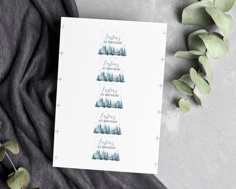 May include: A white rectangular sheet featuring five identical labels arranged vertically. Each label displays the text Easton's 1ST BIRTHDAY above a watercolor illustration of a blue-toned pine forest with a small deer and snowflakes.