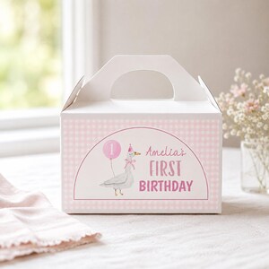 May include: A white cardboard gable box featuring a light pink gingham pattern. The front displays a graphic of a goose wearing a pink party hat and bow, holding a pink balloon with the number 1, and the text Amelia's FIRST BIRTHDAY in pink.