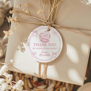 K&ouml;nnte beinhalten: Ein rundes, wei&szlig;es Geschenkanh&auml;nger-Etikett aus Papier mit rosa Tintenillustrationen von Schleifen und einer herzf&ouml;rmigen Torte. Der Text lautet: one, THANK YOU for coming to my party!, Love, Charlotte. Mit einer Schnur an einem Karton befestigt.