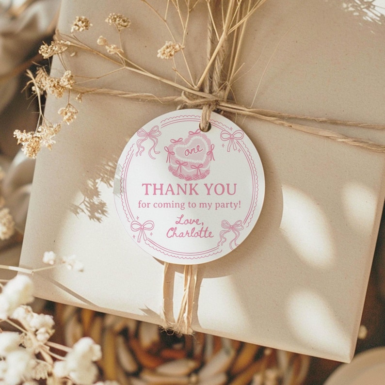 K&ouml;nnte beinhalten: Ein rundes, wei&szlig;es Geschenkanh&auml;nger-Etikett aus Papier mit rosa Tintenillustrationen von Schleifen und einer herzf&ouml;rmigen Torte. Der Text lautet: one, THANK YOU for coming to my party!, Love, Charlotte. Mit einer Schnur an einem Karton befestigt.
