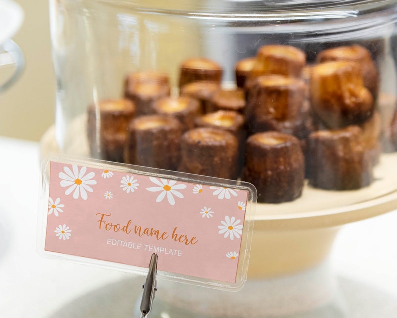 Op de afbeelding: Een doorzichtige glazen stolp over een verzameling kleine, donkerbruine, cilindervormige gebakjes op een beige standaard. Vooraan staat een rechthoekig roze bordje met witte madeliefjes en de tekst Food name here EDITABLE TEMPLATE.