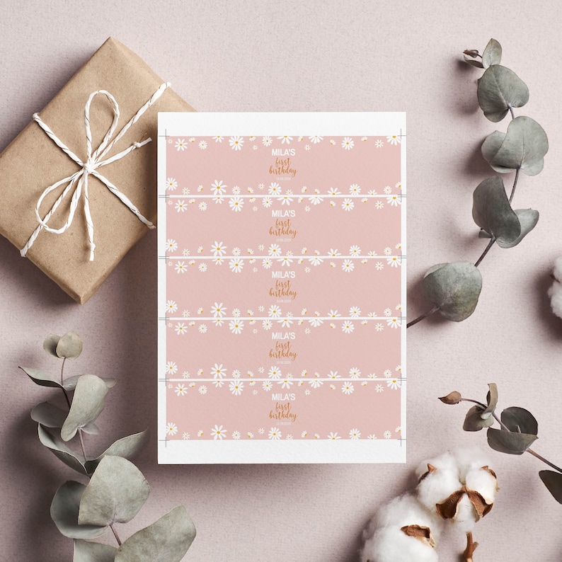 May include: A rectangular sheet of paper featuring five horizontal strips with a dusty pink background, a repeating white daisy pattern, and the gold text "Mila's First Birthday" printed in the center of each strip with crop marks at the edges.