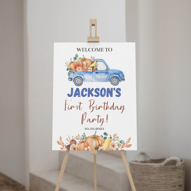 May include: A white rectangular birthday sign on a wooden easel featuring a blue truck illustration carrying pumpkins and flowers. Text reads: WELCOME TO, JACKSON'S, First Birthday Party!, and 10.10.202X. The design includes autumn-themed floral accents.