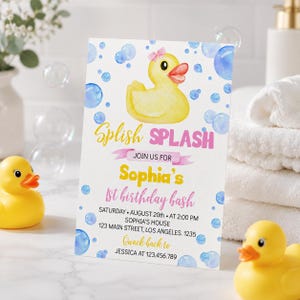 May include: A rectangular birthday invitation card featuring a yellow rubber duck with a pink bow, blue watercolor bubbles, and the text Splish Splash Join us for Sophia's 1st birthday bash Saturday August 20th at 2:00 PM Sophia's house 123 Main Street.