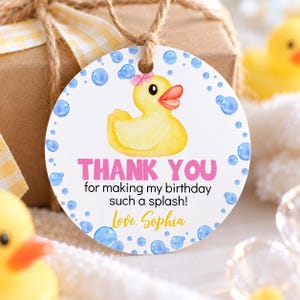 Pu&ograve; includere: Un'etichetta regalo rotonda e bianca con una paperella di gomma gialla con un fiocco rosa, circondata da bolle blu. Il testo recita: THANK YOU for making my birthday such a splash! Love, Sophia. L'etichetta &egrave; fissata con uno spago marrone.