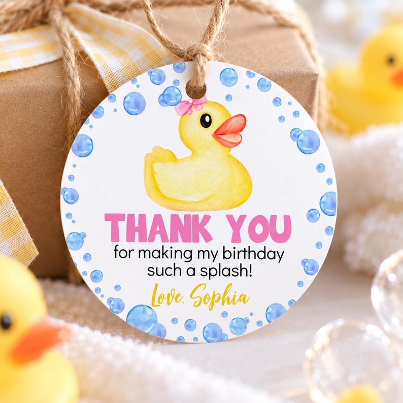 Pu&ograve; includere: Un'etichetta regalo rotonda e bianca con una paperella di gomma gialla con un fiocco rosa, circondata da bolle blu. Il testo recita: THANK YOU for making my birthday such a splash! Love, Sophia. L'etichetta &egrave; fissata con uno spago marrone.