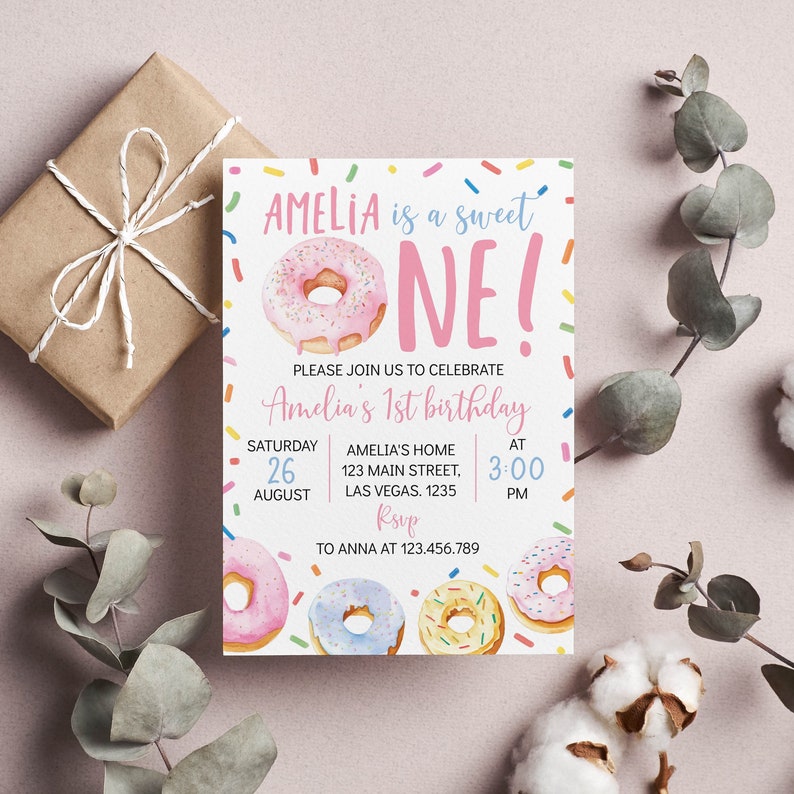 May include: A rectangular birthday invitation card featuring a pink frosted donut illustration and the text AMELIA is a sweet ONE! PLEASE JOIN US TO CELEBRATE Amelia's 1st birthday SATURDAY 26 AUGUST AMELIA'S HOME 123 MAIN STREET LAS VEGAS 1235 AT 3:00 PM RSVP TO ANNA AT 123.456.789.