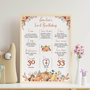 May include: A rectangular framed milestone poster titled Amelia's First Birthday with autumn-themed floral and pumpkin illustrations. The white sign lists personal details including I can, I say, Nicknames, I weigh 30 pounds, 4 teeth, and 33 inches.