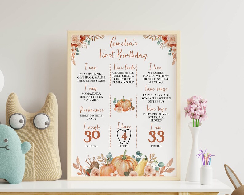 May include: A rectangular framed milestone poster titled Amelia's First Birthday with autumn-themed floral and pumpkin illustrations. The white sign lists personal details including I can, I say, Nicknames, I weigh 30 pounds, 4 teeth, and 33 inches.
