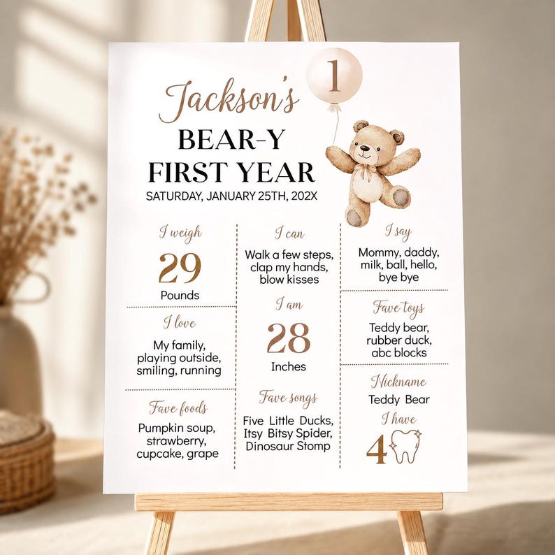 May include: A white milestone birthday sign titled Jackson's BEAR-Y FIRST YEAR with a brown teddy bear illustration holding a balloon with the number 1. The sign lists developmental milestones, weight in pounds, height in inches, and favorite items.