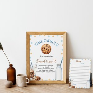 Op de afbeelding: Een ingelijst bord met blauwe en gouden stippen waarop "Time Capsule" staat met een afbeelding van een chocolade chip cookie. Het bord zegt "To be opened when Daniel turns 18". Er is een kleiner kaartje waarop "Dear Daniel" staat met regels om een ​​bericht te schrijven.