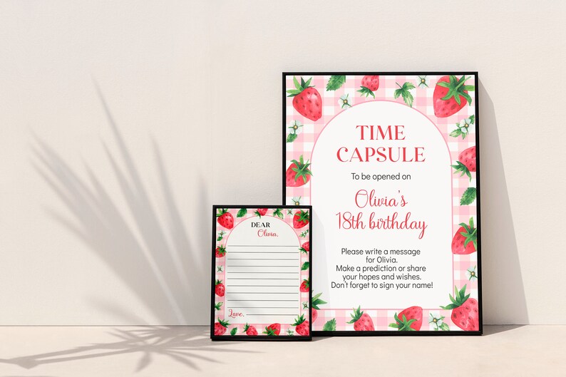 May include: Two framed signs with a pink and white gingham background and watercolor strawberry illustrations. The larger sign reads TIME CAPSULE To be opened on Olivia's 18th birthday. The smaller sign has lines for messages and reads DEAR Olivia.