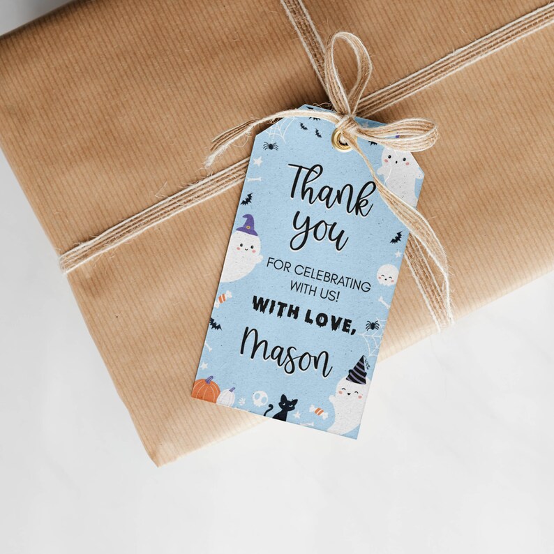 Pu&ograve; includere: Un'etichetta regalo rettangolare azzurra con un occhiello dorato e un fiocco in spago naturale. L'etichetta presenta illustrazioni a tema Halloween con fantasmi, zucche, un gatto nero e ragni, con il testo Thank you FOR CELEBRATING WITH US! WITH LOVE, MASON.