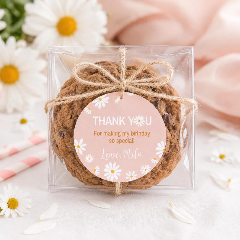 May include: A chocolate chip cookie packaged in a clear square box, tied with twine and a round pink tag featuring white daisy illustrations. The tag reads: THANK YOU For making my birthday so special! Love, Mila, with a daisy replacing the letter O.
