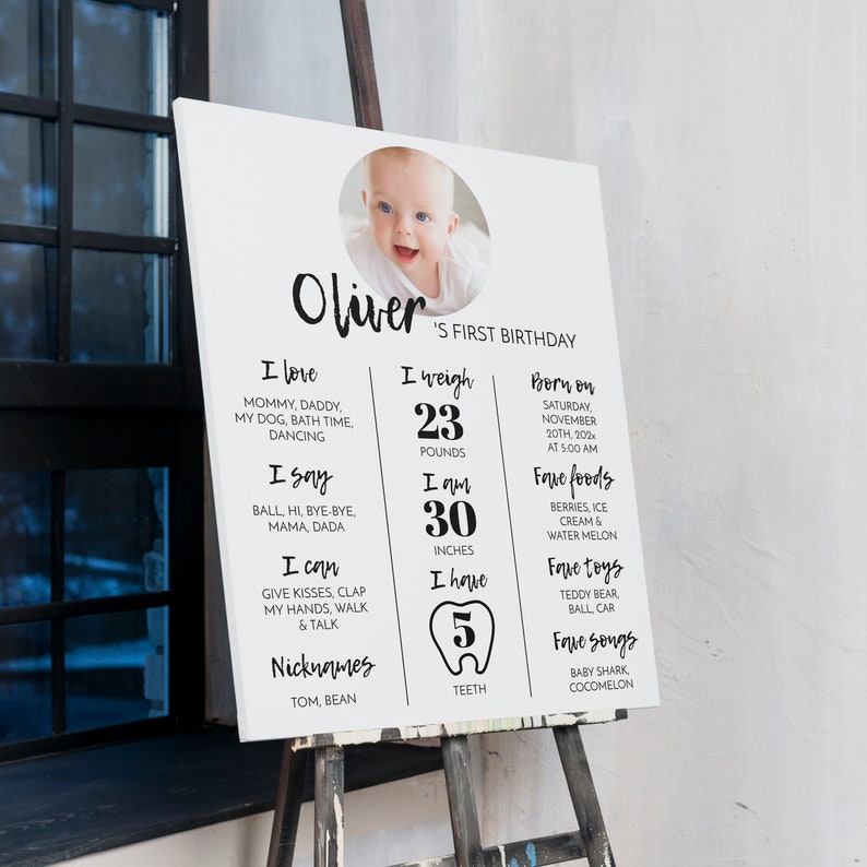 May include: A white rectangular milestone birthday sign featuring a circular photo of a baby at the top. The text reads Oliver's First Birthday, listing personal details including weight in pounds, height in inches, and five teeth count.