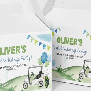 K&ouml;nnte beinhalten: Zwei wei&szlig;e Pappschachteln mit einer Aquarellillustration eines Golfwagens auf einem gr&uuml;nen H&uuml;gel, blauen und gr&uuml;nen Luftballons und Wimpeln. Der Text lautet: OLIVER'S First Birthday Party! THANK YOU FOR CELEBRATING WITH ME!