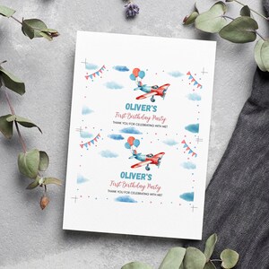 May include: A white rectangular card featuring two identical prints of a red and blue airplane illustration with balloons, blue clouds, and bunting. The text reads: OLIVER'S First Birthday Party THANK YOU FOR CELEBRATING WITH ME! with crop marks.
