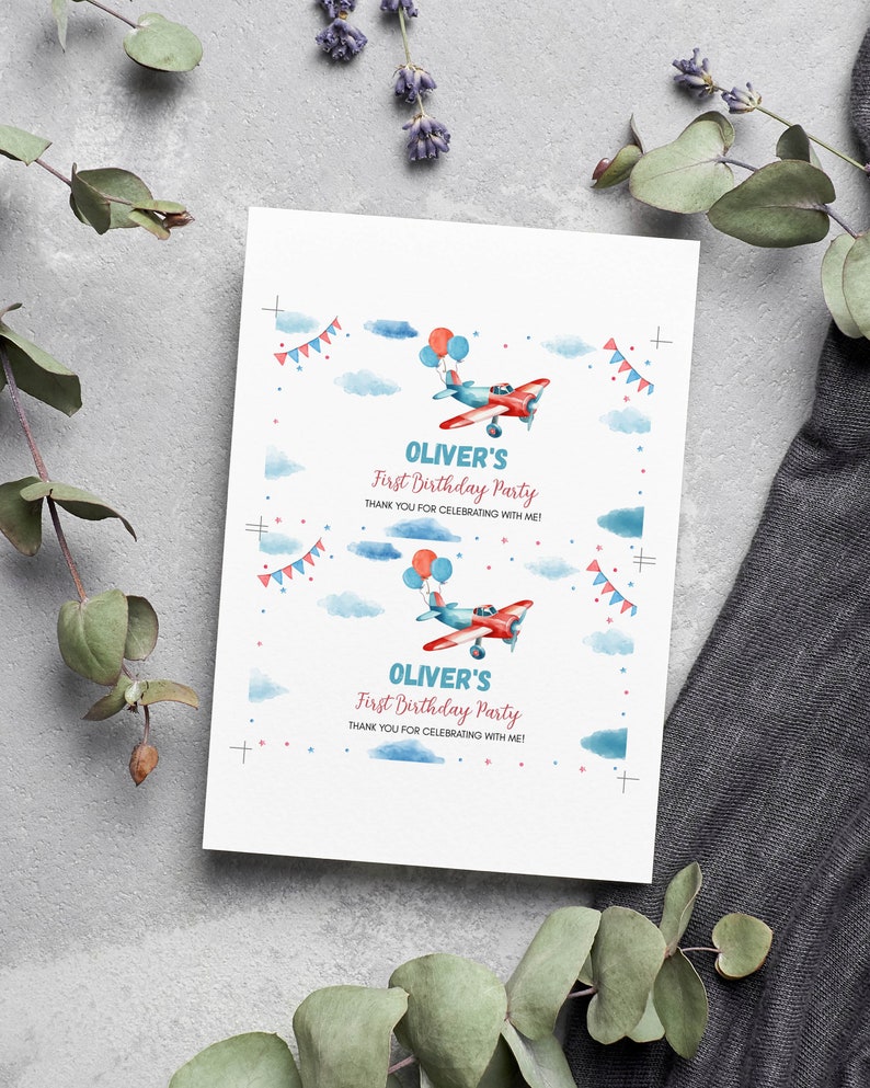 May include: A white rectangular card featuring two identical prints of a red and blue airplane illustration with balloons, blue clouds, and bunting. The text reads: OLIVER'S First Birthday Party THANK YOU FOR CELEBRATING WITH ME! with crop marks.