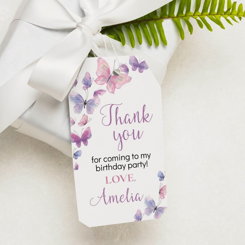 May include: A white rectangular gift tag with clipped corners featuring watercolor butterflies in shades of pink and purple. The text reads: Thank you for coming to my birthday party! LOVE, Amelia. The tag is attached to a white gift box.