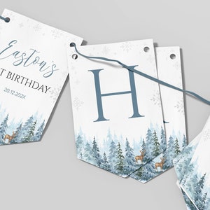 Puede incluir: Una guirnalda decorativa de papel con tem&aacute;tica de bosque invernal, pinos azules, ciervos y copos de nieve. Los banderines muestran el texto Easton's 1ST BIRTHDAY 20.12.202X y la letra H, unidos por un cord&oacute;n azul para colgar.