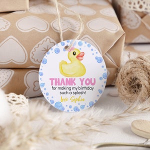 Pu&ograve; includere: Un'etichetta regalo rotonda bianca con una papera di gomma gialla con un fiocco rosa, bolle blu e il testo THANK YOU for making my birthday such a splash! Love Sophia. L'etichetta &egrave; sospesa da un sottile spago di juta per i regali.