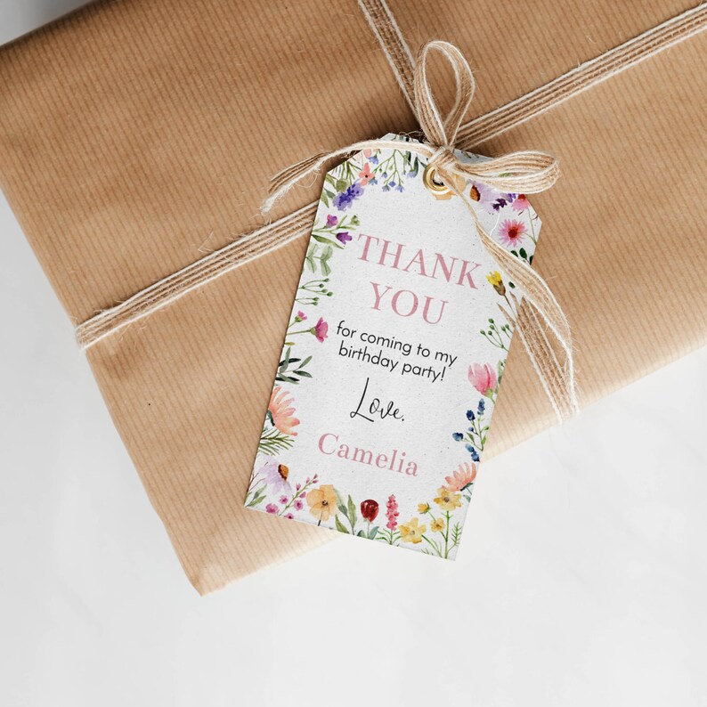 May include: A rectangular paper gift tag with a floral border featuring pink, yellow, blue, and green wildflowers. The text reads: THANK YOU for coming to my birthday party! Love. Camelia. The tag has a gold eyelet and is tied with a tan twine bow.