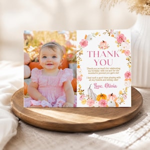May include: A rectangular thank you card featuring a photo of a smiling baby in a pink dress on the left and a floral wreath with pumpkins on the right. The text reads: THANK YOU, Thank you so much for celebrating my birthday with me and for the wonderful present you gave me! I had such a good time playing with all my friends and eating cake! Love, Olivia.