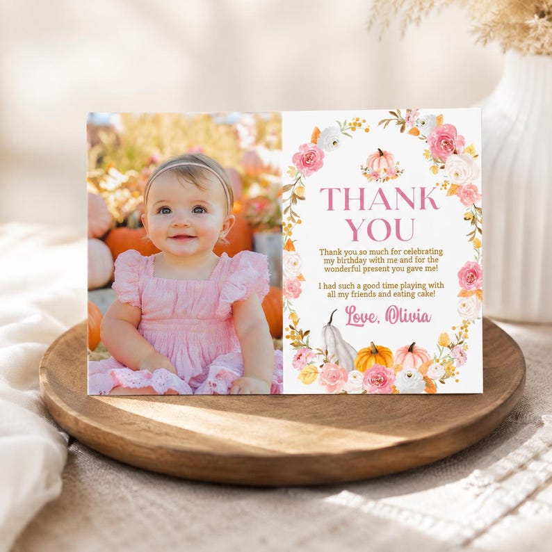 May include: A rectangular thank you card featuring a photo of a smiling baby in a pink dress on the left and a floral wreath with pumpkins on the right. The text reads: THANK YOU, Thank you so much for celebrating my birthday with me and for the wonderful present you gave me! I had such a good time playing with all my friends and eating cake! Love, Olivia.