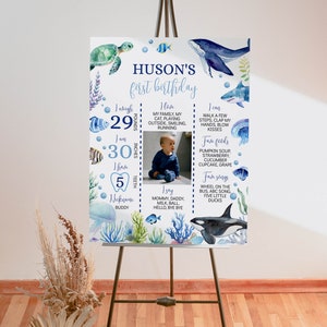 May include: A blue and white first birthday sign with a watercolor ocean theme. The sign includes a photo of a baby, the baby's name "Huson's", and details about the baby's first year, including weight, height, teeth, favorite foods, and songs.