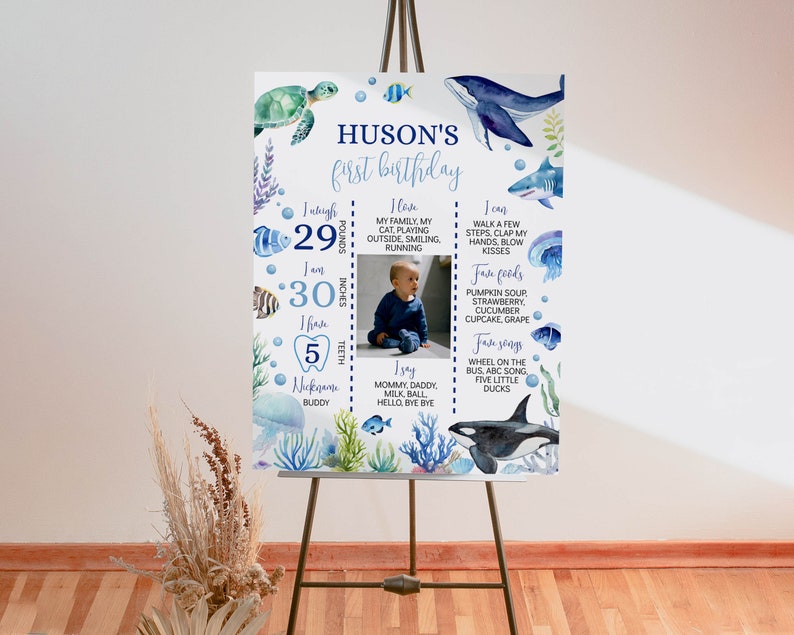 May include: A blue and white first birthday sign with a watercolor ocean theme. The sign includes a photo of a baby, the baby's name "Huson's", and details about the baby's first year, including weight, height, teeth, favorite foods, and songs.