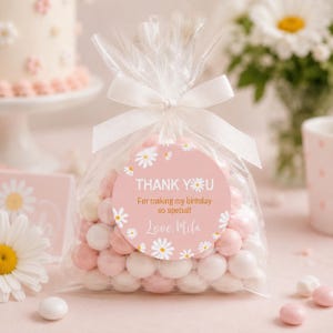 May include: A clear plastic favor bag filled with round white and light pink candies, tied with a white ribbon. A circular sticker on the front features a pink background with white daisy illustrations and the text: THANK YOU For making my birthday so special! Love, Mila.