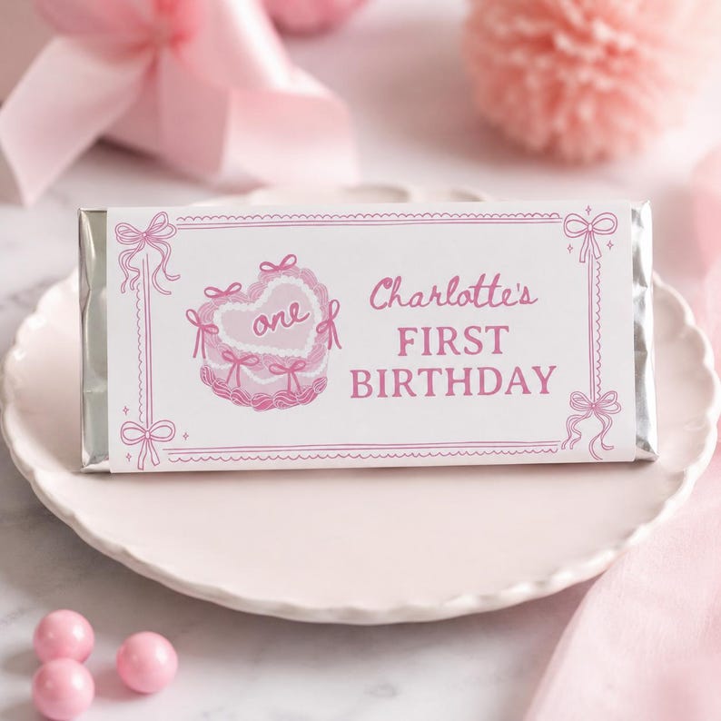 May include: A rectangular chocolate bar wrapper featuring a white background with pink decorative borders and bows. The design includes a pink heart-shaped cake illustration with the word one and the text Charlotte's FIRST BIRTHDAY in pink.