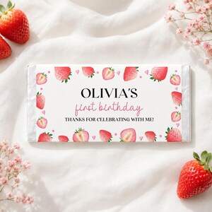 May include: A rectangular candy bar wrapper featuring a white background with a border of watercolor strawberries and small pink hearts. The text reads OLIVIA'S first birthday THANKS FOR CELEBRATING WITH ME! in black and pink lettering.