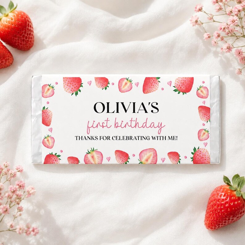 May include: A rectangular candy bar wrapper featuring a white background with a border of watercolor strawberries and small pink hearts. The text reads OLIVIA'S first birthday THANKS FOR CELEBRATING WITH ME! in black and pink lettering.