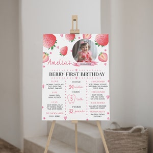 Op de afbeelding: Een wit verjaardagsbord met aquarel aardbeien, een ronde kinderfoto en tekst waaronder Amelia's Berry First Birthday, 86 cm, 5 tanden en 15 kg, weergegeven op een houten ezel voor een feestelijke gelegenheid.