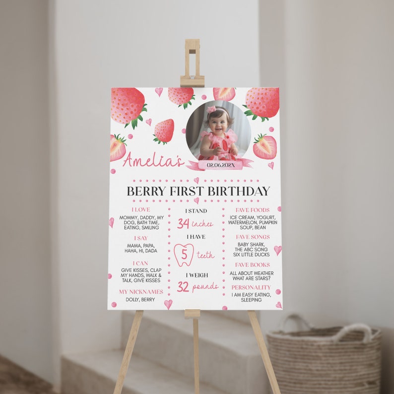 Op de afbeelding: Een wit verjaardagsbord met aquarel aardbeien, een ronde kinderfoto en tekst waaronder Amelia's Berry First Birthday, 86 cm, 5 tanden en 15 kg, weergegeven op een houten ezel voor een feestelijke gelegenheid.