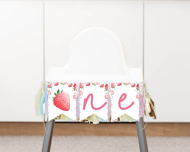 Puede incluir: Una guirnalda para trona con tres banderines blancos decorados con ilustraciones de fresas en acuarela y las letras cursivas rosas O, N y E. La guirnalda est&aacute; decorada con borlas de tela colgantes en tonos pastel a ambos lados.