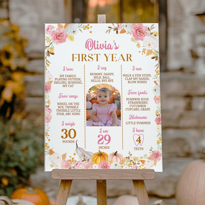 May include: A white milestone birthday sign featuring pink and white floral borders, gold text, and a central photograph of a child. The sign displays text including Olivia's First Year, I love, I say, I can, Fave foods, Fave songs, and I weigh 30 pounds.