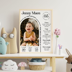Puede incluir: Un p&oacute;ster personalizado para el primer cumplea&ntilde;os con una foto de una ni&ntilde;a, que incluye su nombre, fecha de nacimiento, peso, altura y cosas favoritas. El p&oacute;ster tiene un fondo de madera clara y un arco blanco con texto negro.