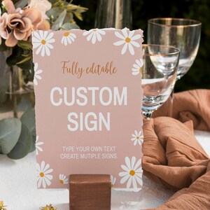 May include: A rectangular sign with a dusty rose background and white daisy illustrations along the edges. The text reads Fully editable CUSTOM SIGN and TYPE YOUR OWN TEXT CREATE MUTIPLE SIGNS. The sign is held upright by a small brown wooden block.