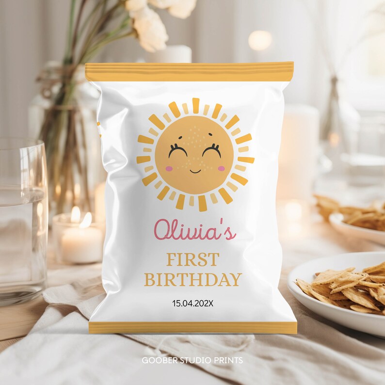 May include: A white chip bag with a yellow border and a smiling sun design. The text "Olivia's First Birthday 15.04.202X" is printed on the bag.