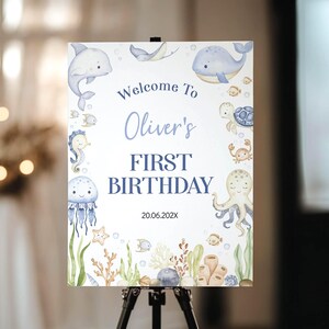 May include: A rectangular sign featuring watercolour illustrations of sea creatures including a dolphin, whale, turtle, crab, octopus, and jellyfish. The text reads Welcome To Oliver's FIRST BIRTHDAY 20.06.202X, surrounded by coral and seaweed.