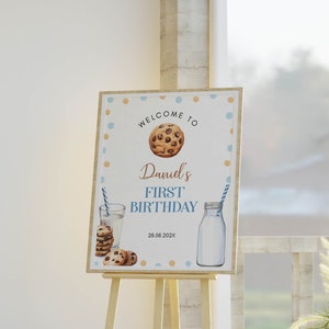 May include: Welcome to Daniel's First Birthday sign with a blue and brown polka dot background. The sign features a chocolate chip cookie, a glass of milk with a straw, and a bottle of milk. The date 28.08.202X is printed on the sign.
