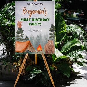 Op de afbeelding: Een rechthoekig bord op een houten ezel met de tekst WELCOME TO Benjamin's FIRST BIRTHDAY PARTY! 20.10.202X. Het ontwerp bevat een aquarelillustratie van een bruine beer, een eekhoorn, een tent, een kampvuur en dennenbomen.