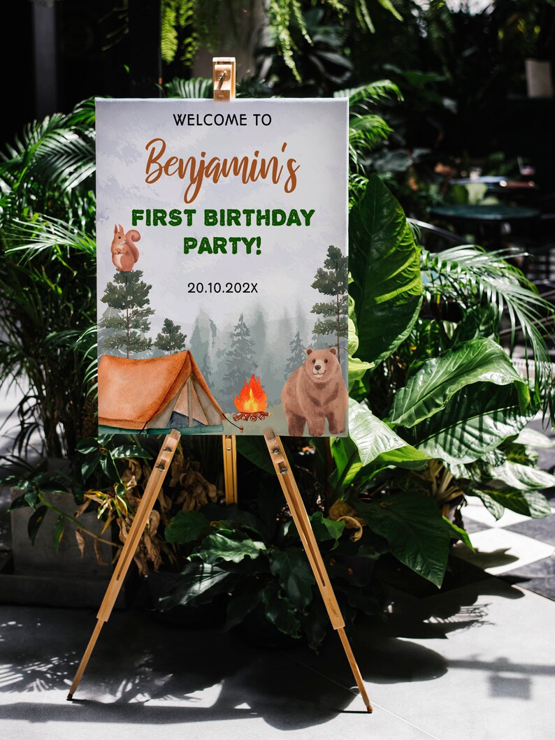 Op de afbeelding: Een rechthoekig bord op een houten ezel met de tekst WELCOME TO Benjamin's FIRST BIRTHDAY PARTY! 20.10.202X. Het ontwerp bevat een aquarelillustratie van een bruine beer, een eekhoorn, een tent, een kampvuur en dennenbomen.