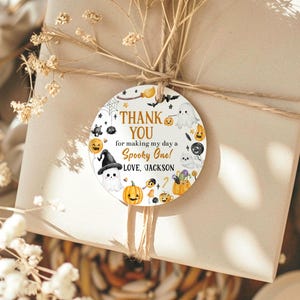May include: A round Halloween-themed gift tag with the text "THANK YOU for making my day a Spooky One! LOVE, JACKSON." The tag features illustrations of ghosts, pumpkins, bats, and a witch's hat. The tag is tied to a wrapped gift with twine.